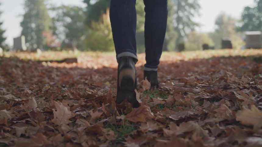 A woman with boots walking on leaves in the cemetery with trees and the sky in the background