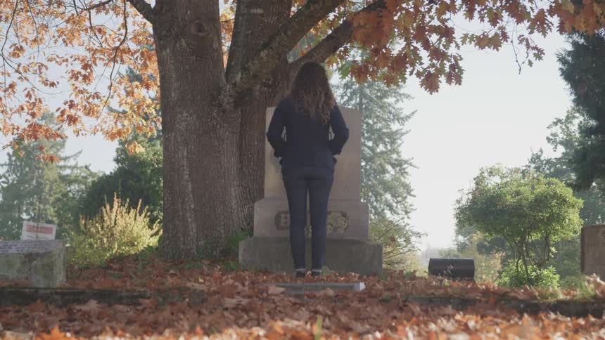 A woman stands in front of a gravestone at a cemetery