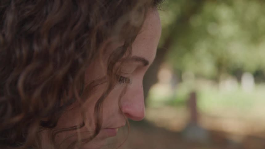 A woman with brown curly hair is in a cemetery looking down in deep thought