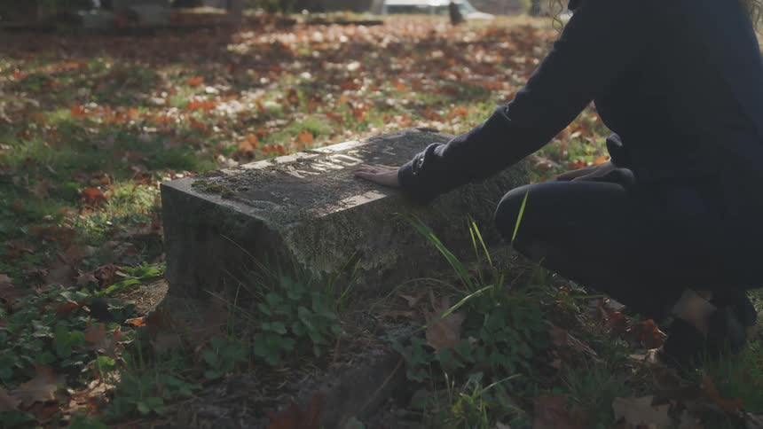 A woman kneels down in a cemetery and brushes dirt off a tombstone
