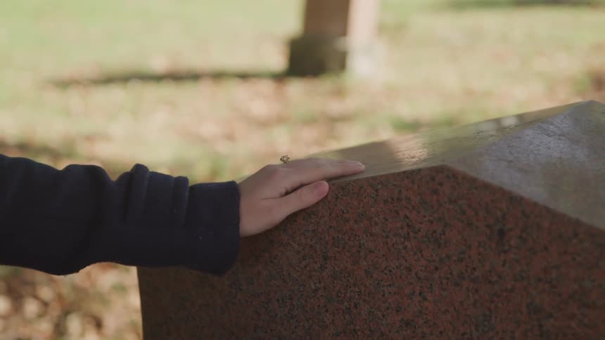 A woman stands with her hand upon a gravestone