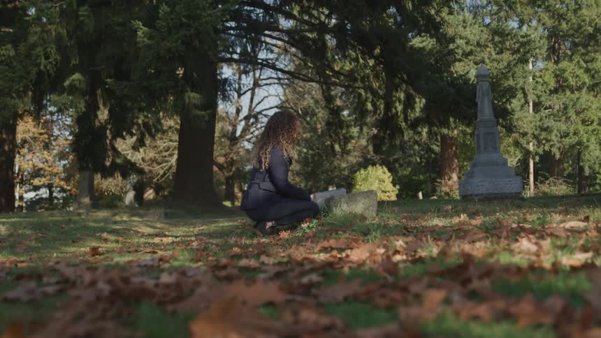 A woman kneeling at a headstone in a cemetery