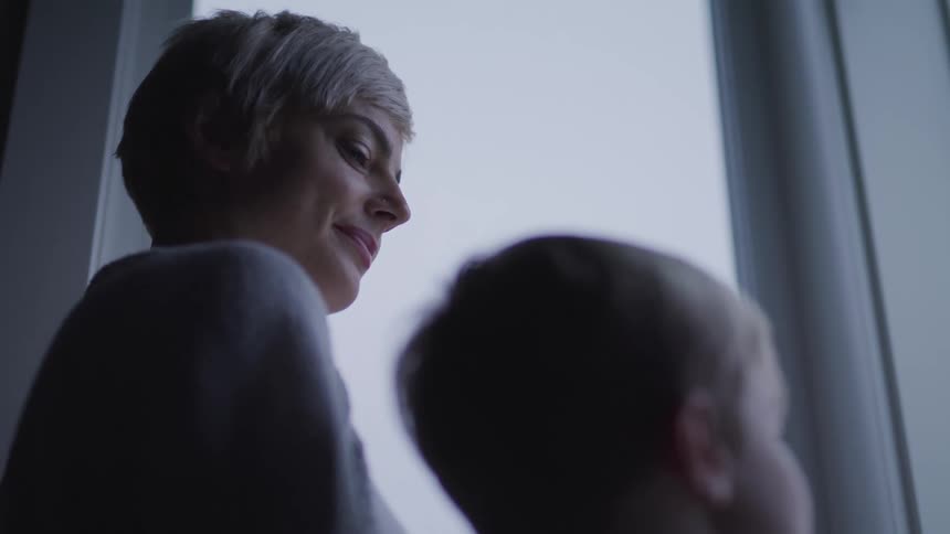 Mother with a short blonde hair stands and looks at her son looking out the window