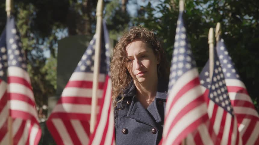 A woman stands solemnly before a grave covered in american flags in a cemetery during the day