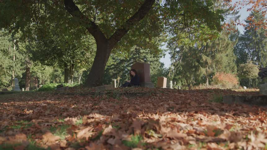 A woman sitting on the ground and leaning against a headstone in a cemetery