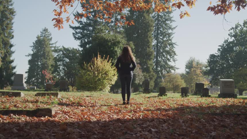 A woman stands motionlessly among the headstones in a cemetery during the day