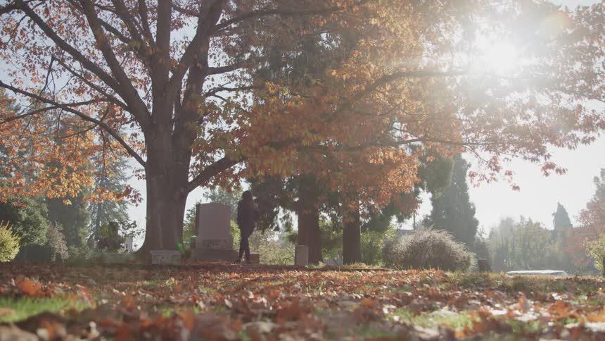 A white woman with long hairin a sweater and pants walks through a cemetery filled with leaved and gravestones going to some ones grave