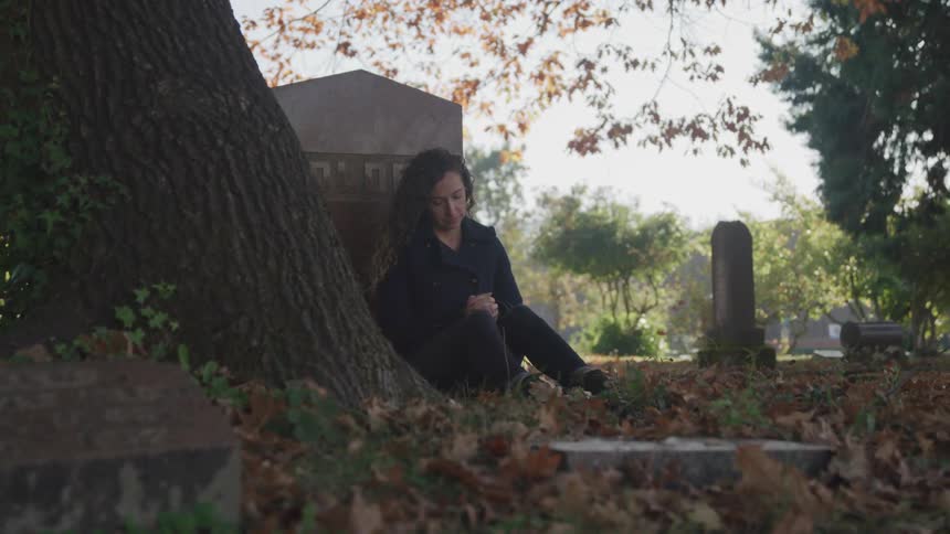 A young woman prays as she leans against a headstone in a cemetery