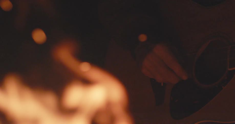 A person strums an acoustic guitar next to an open fire at night
