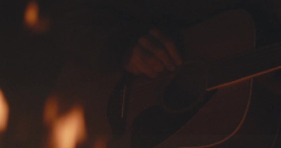 Man sits in front of a campfire with a guitar in his lap at night