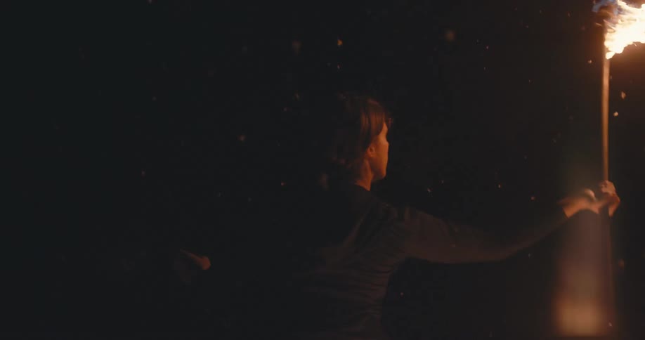 Person stands outside at night spinning fire torch in falling snow