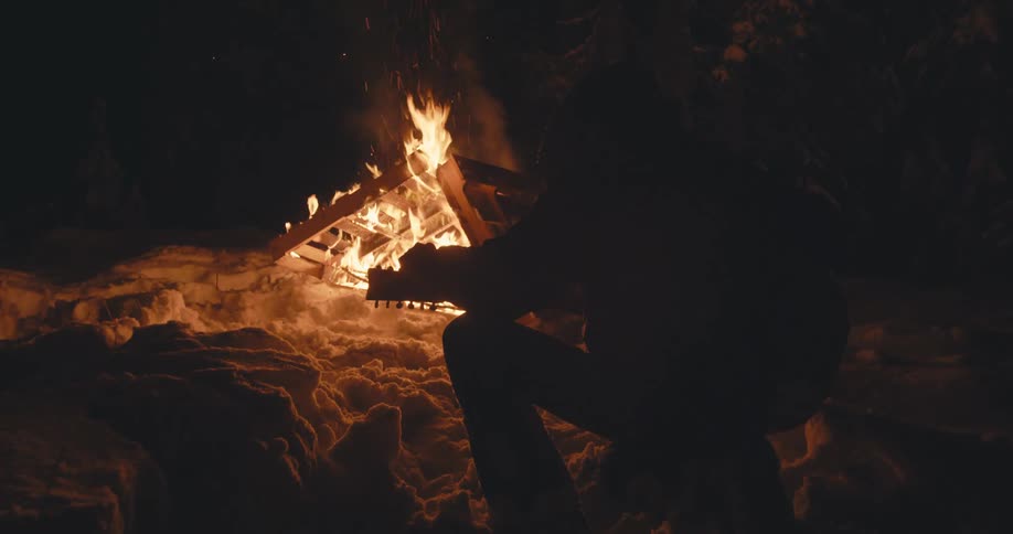 A person sits near a small bonfire in the snow as they tune their guitar