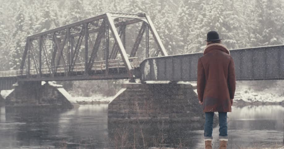 A man stands by a river bridge with light snow falling around him