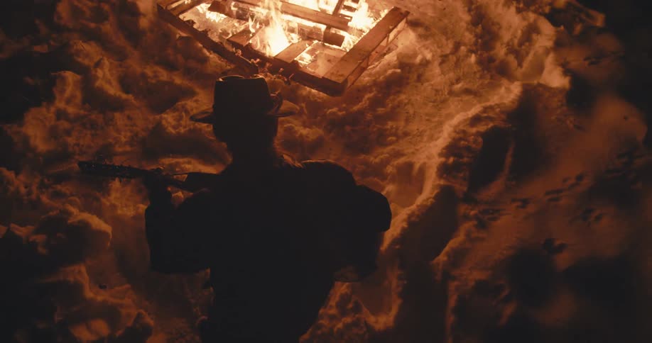 A guitarist plays the guitar while standing outdoors in the snow near a campfire at night