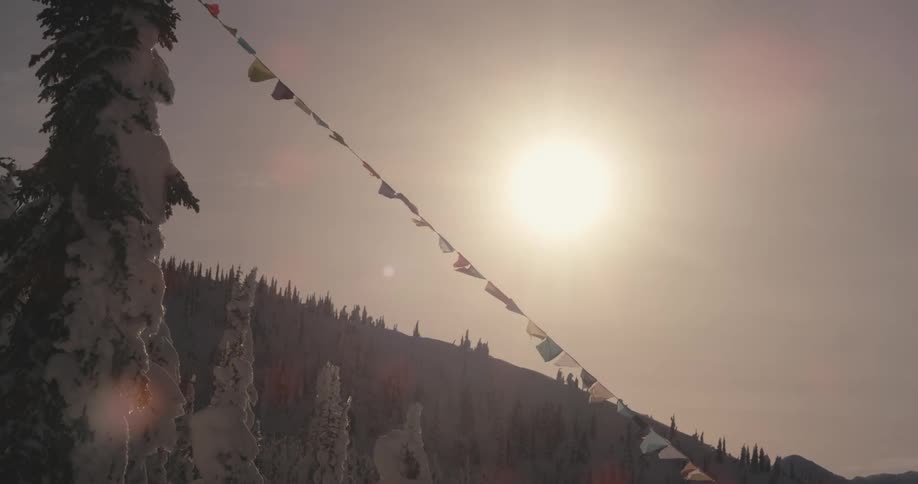 The sun rises over a snowy mountain range where prayer flags fly from a tree