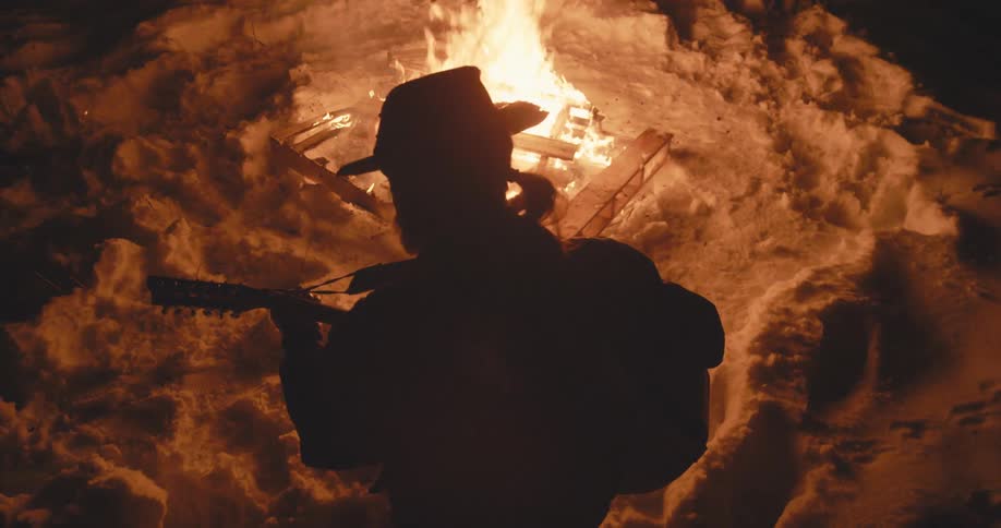 Silhouette of man holding guitar sways standing in area lit by campfire