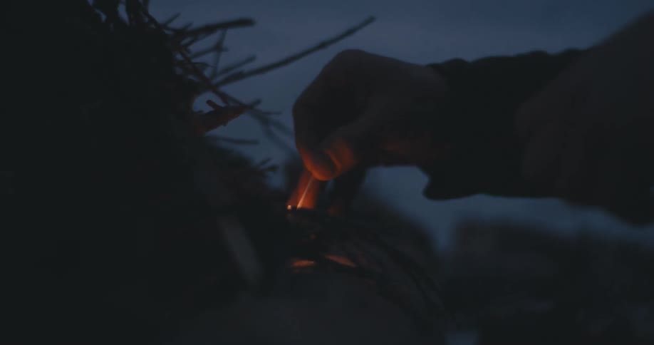 Man wearing coat strikes match and lights pile of wood and kindling outdoors at night