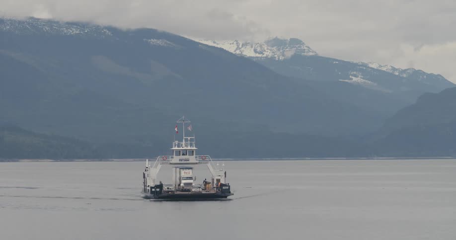 Ferry carries semi through water passing by mountain with snow covered peak on a misty day