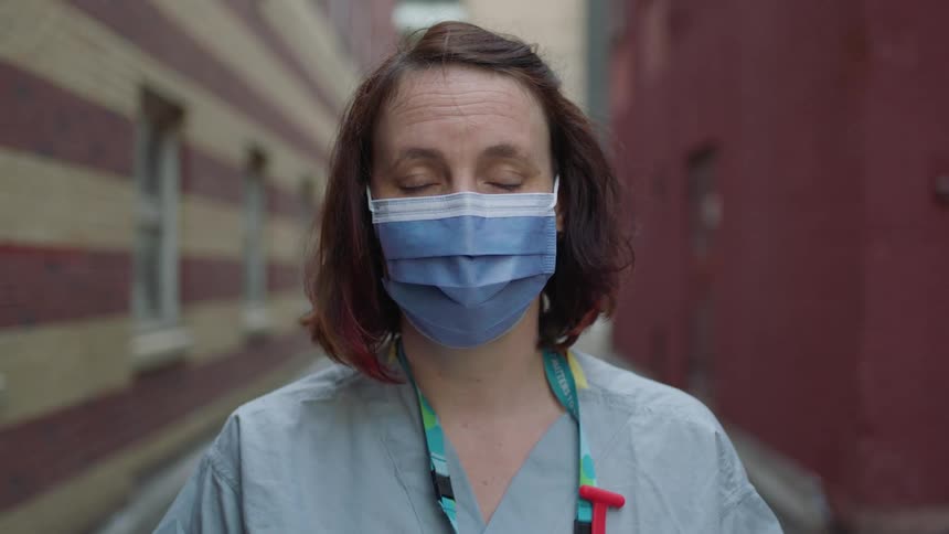A white female medical worker stands outdoors and stares