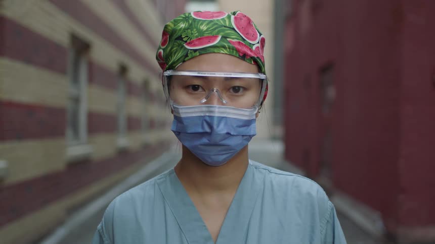 A nurse stands in an alley wearing goggles and a surgical mask