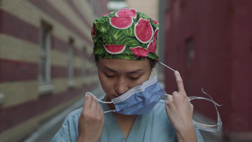 A nurse in scrubs removes a surgical mask in an alley