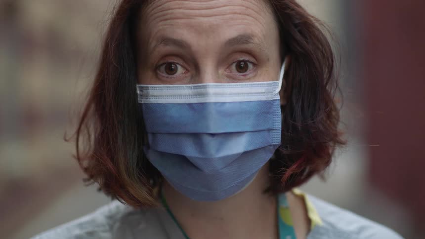 A white female medical worker wearing a face mask stands outdoors and stares