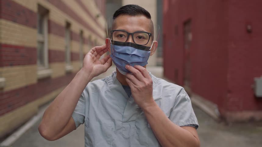 A man in scrubs wearing a mask and face shield standing in the alley behind a hospital building takes off his mask