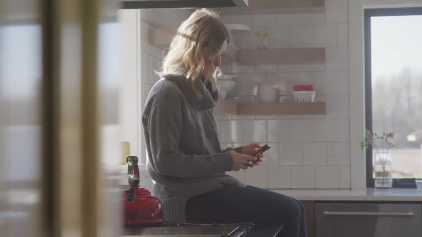 A woman is sitting on the kitchen counter next to a stove while she uses her cell phone