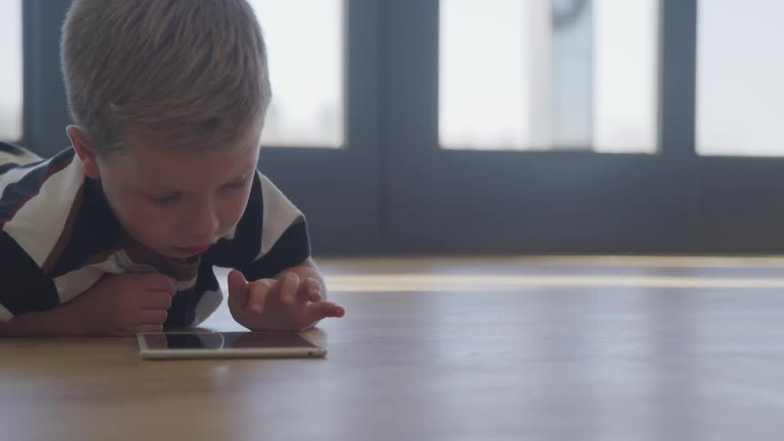 A toddler boy lays on a hardwood floor looking at a tablet