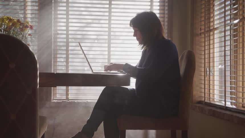 Woman sitting in a table at home types on a laptop near a window