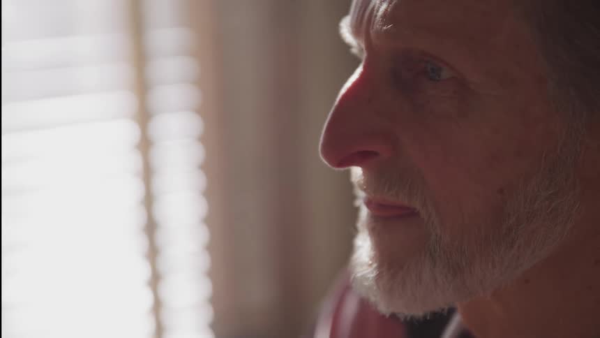 Elderly man licks lips as sunlight comes through window blinds