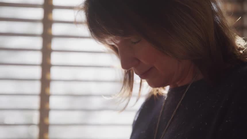 A woman stands in her living room with her head bowed and her eyes closed