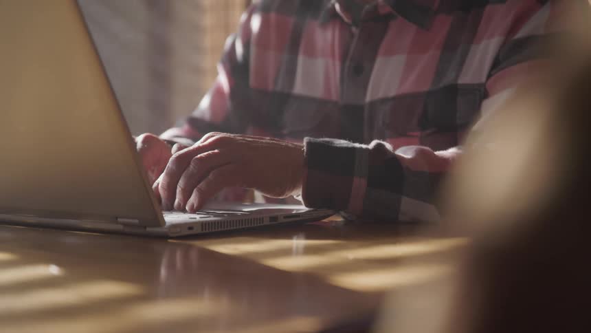Man with a plaid shirt sits at table and uses grey laptop