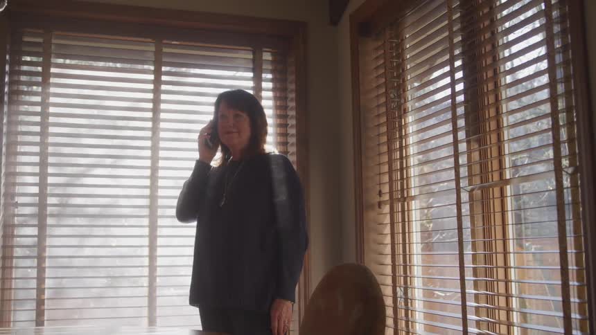 A woman standing next to a chair while talking on the phone with windows in the background