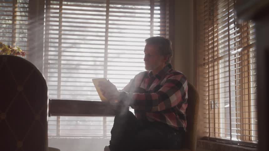 Man sits at table in corner bordered by two windows reading on tablet