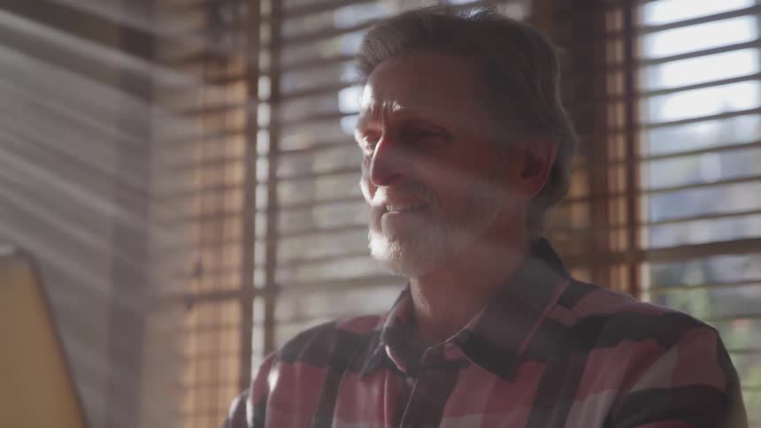 A man sitting looking at a laptop with windows and blinds in the background