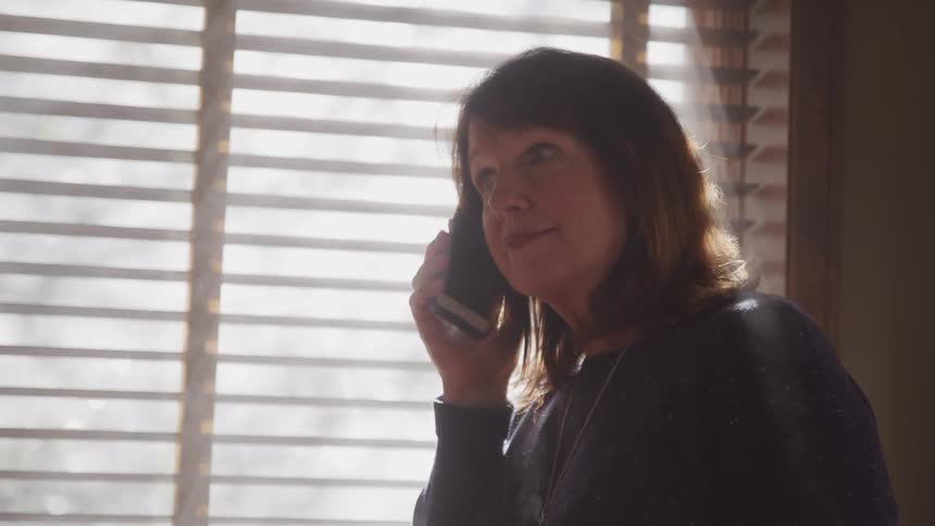 A woman sits by a large window talking on a cell phone