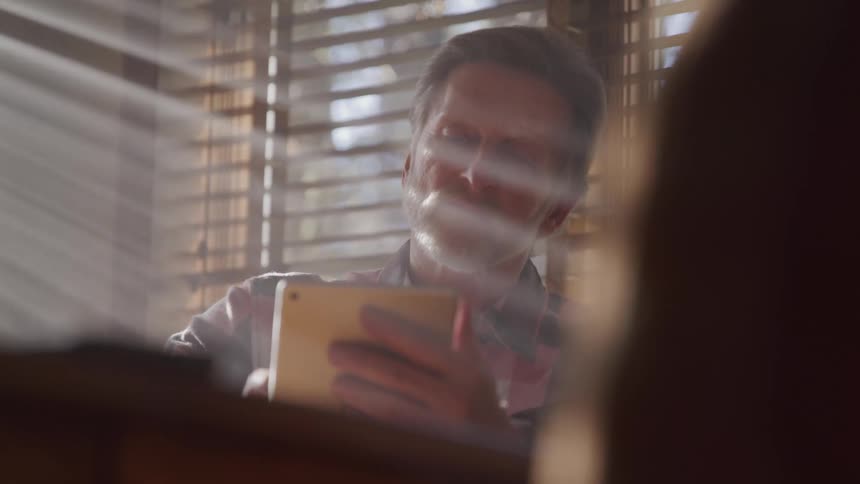 Elderly man with a beard and a plaid shirt uses a tablet as the sun shines through windows in a room