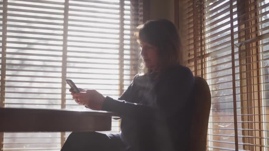 A woman sitting on a chair next to a table holding a smart phone with windows in the background