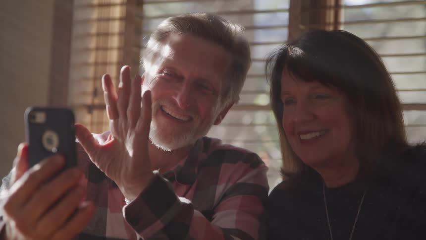 Male holds cellphone while he and his wife smile and wave while video chatting in front of a window indoors