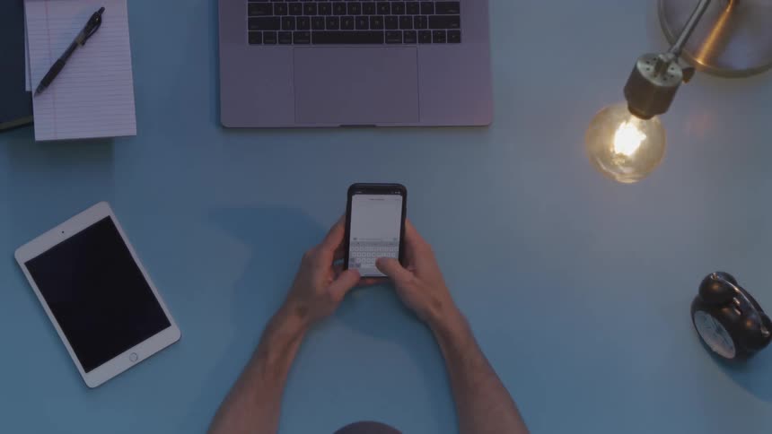A person holds up their smart phone and quickly types a message before setting the phone down near a computer and tablet