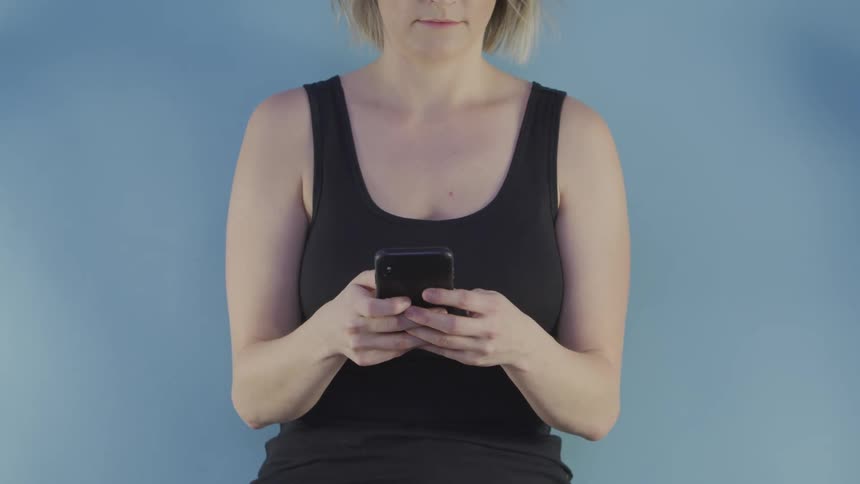 Blonde woman with a black tank top uses a smartphone in front of a blue wall