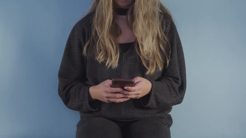 Blonde woman sits against blue background dialing on mobile phone then raises phone to ear