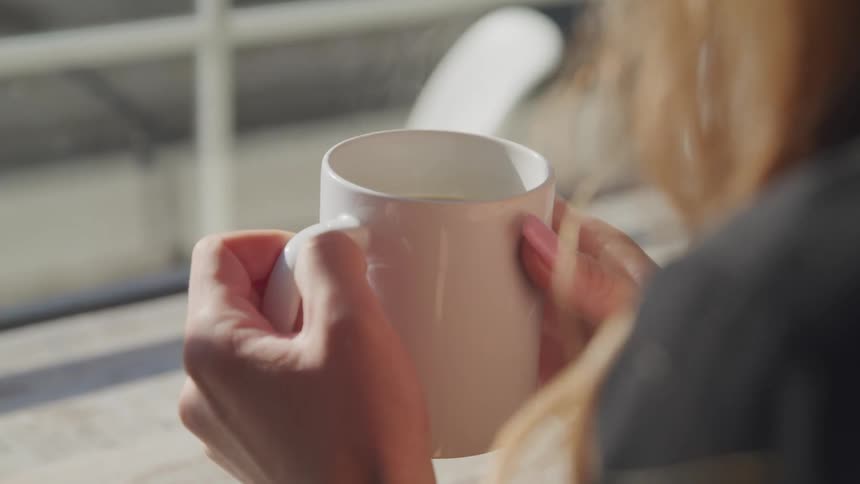 A woman sits by a window and holds a mug of a hot liquid