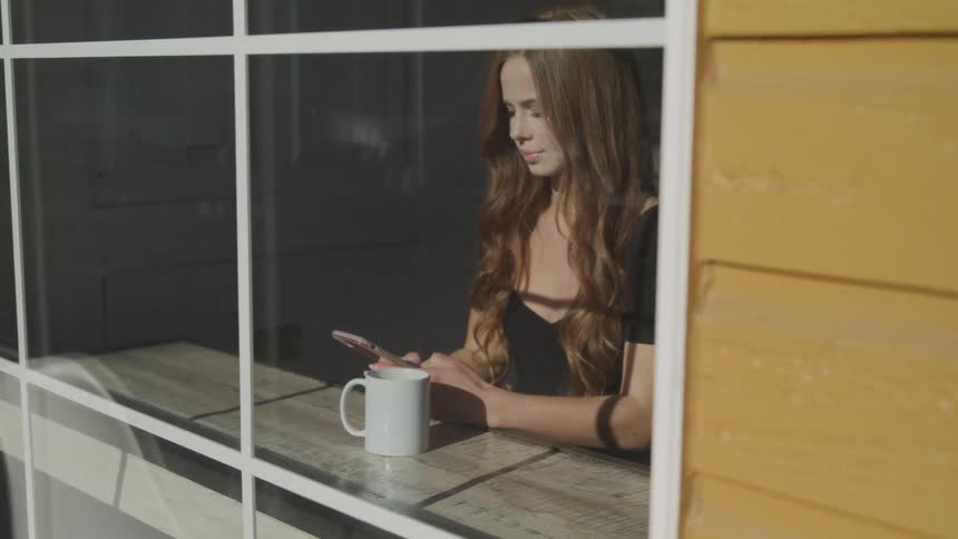 A woman scrolls on her smartphone while seated at on a counter facing a window in a coffee shop