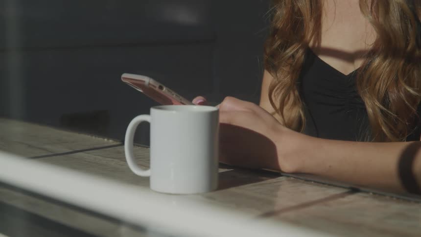 Sunlight shines through a window onto a woman typing on her smartphone while sitting at a counter in front a coffee mug