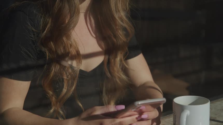 A young girl in a cafe sits at a table with a cup of coffee and scrolls on her phone