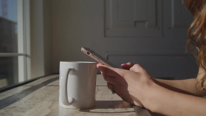 A woman types on a smartphone while sitting at a counter with a coffee cup