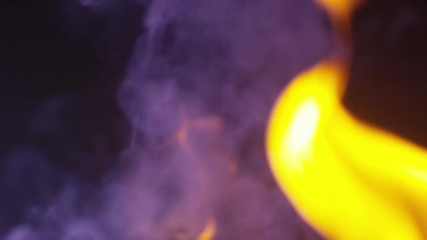 Flame is shades of yellow burns against dark background as purple shaded smoke rises