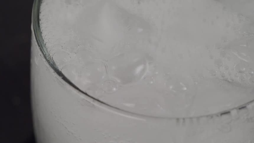 Liquid is poured into a clear glass with ice cubes creating bubbles and fizz and foam